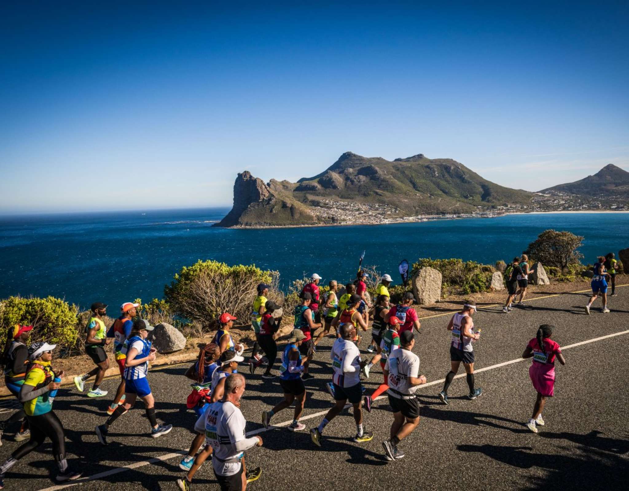 Des marathoniens courent sur une route côtière avec vue sur la mer et des montagnes en arrière-plan