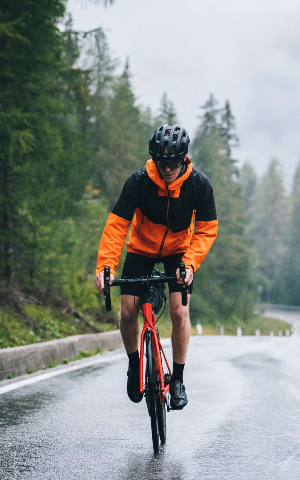 Cycliste en veste de pluie noire et orange roulant sous la pluie sur une route mouillée bordée de conifères.