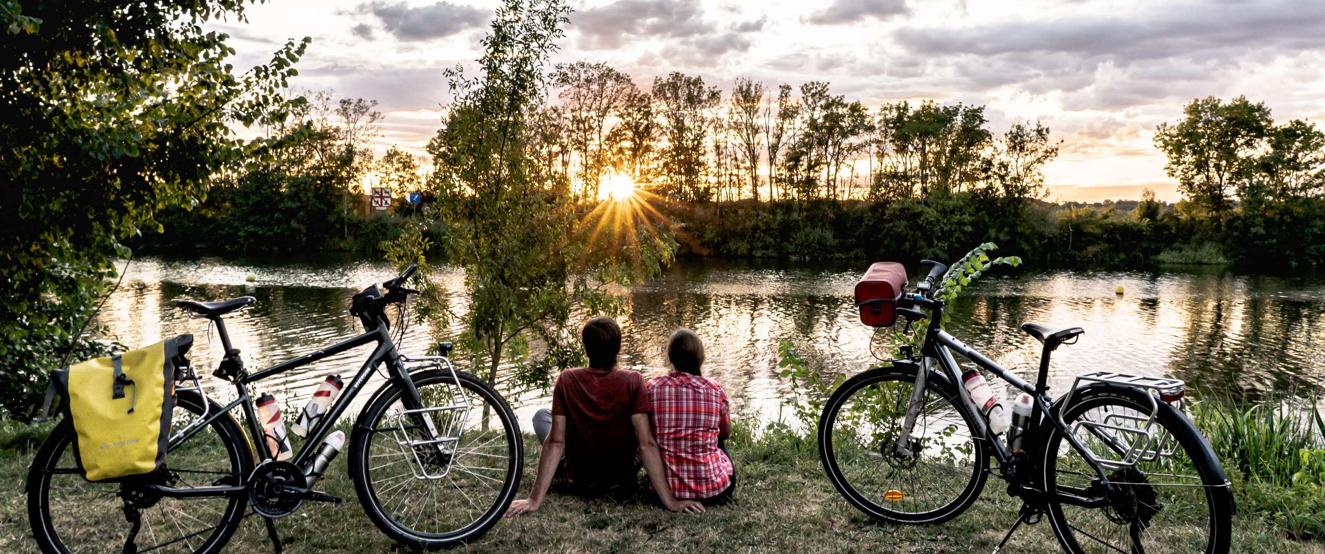 Deux vélos sont garés au bord de l’eau pendant que deux personnes regardent le coucher du soleil.