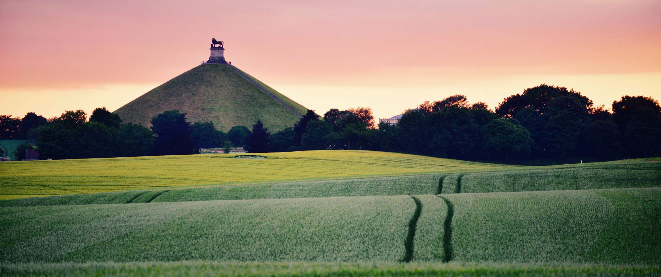 Paysage de champs, colline verte et statue sous un ciel coloré.