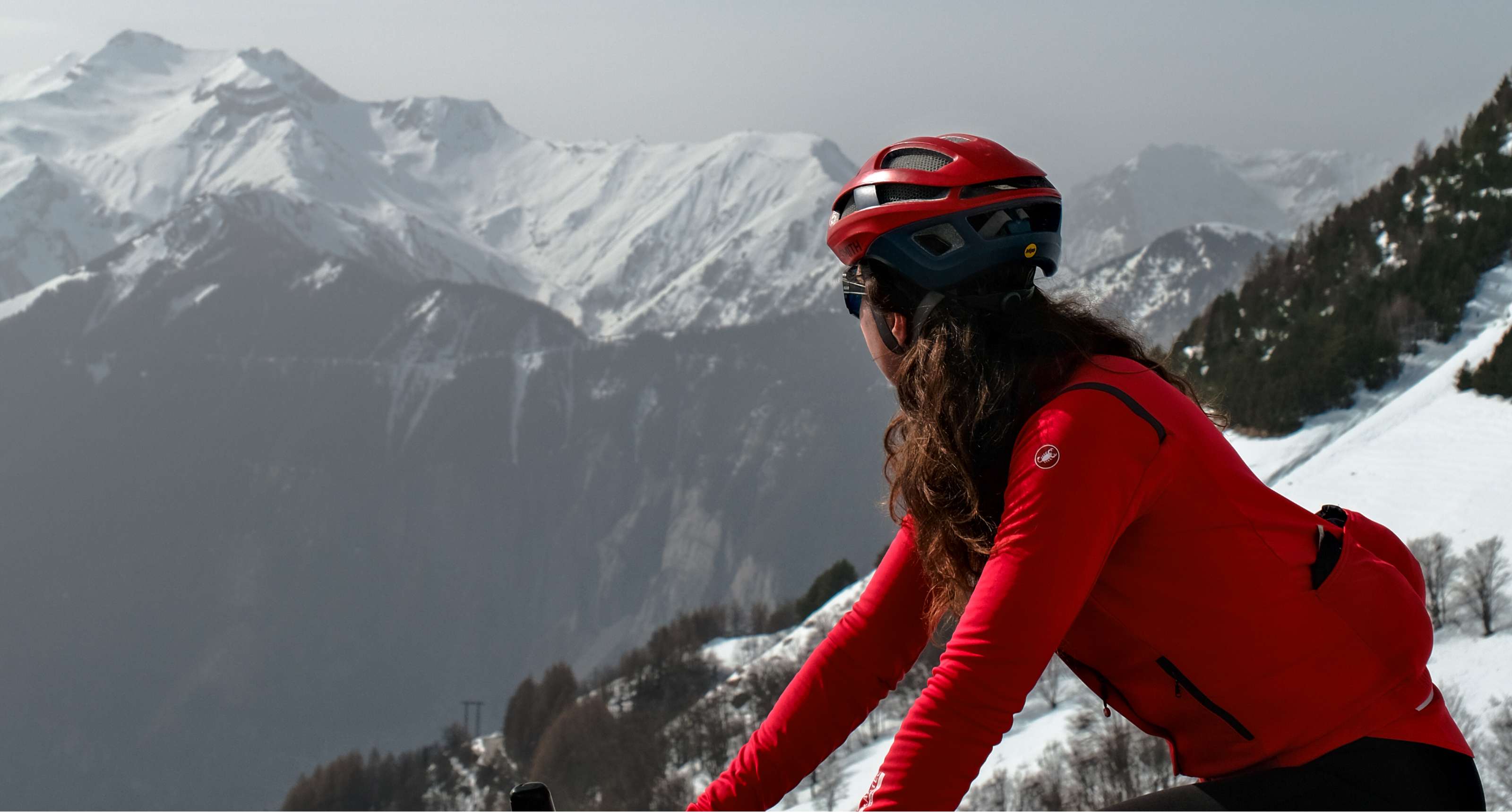Une cycliste avec un casque rouge admire un paysage montagneux aux sommets enneigés.