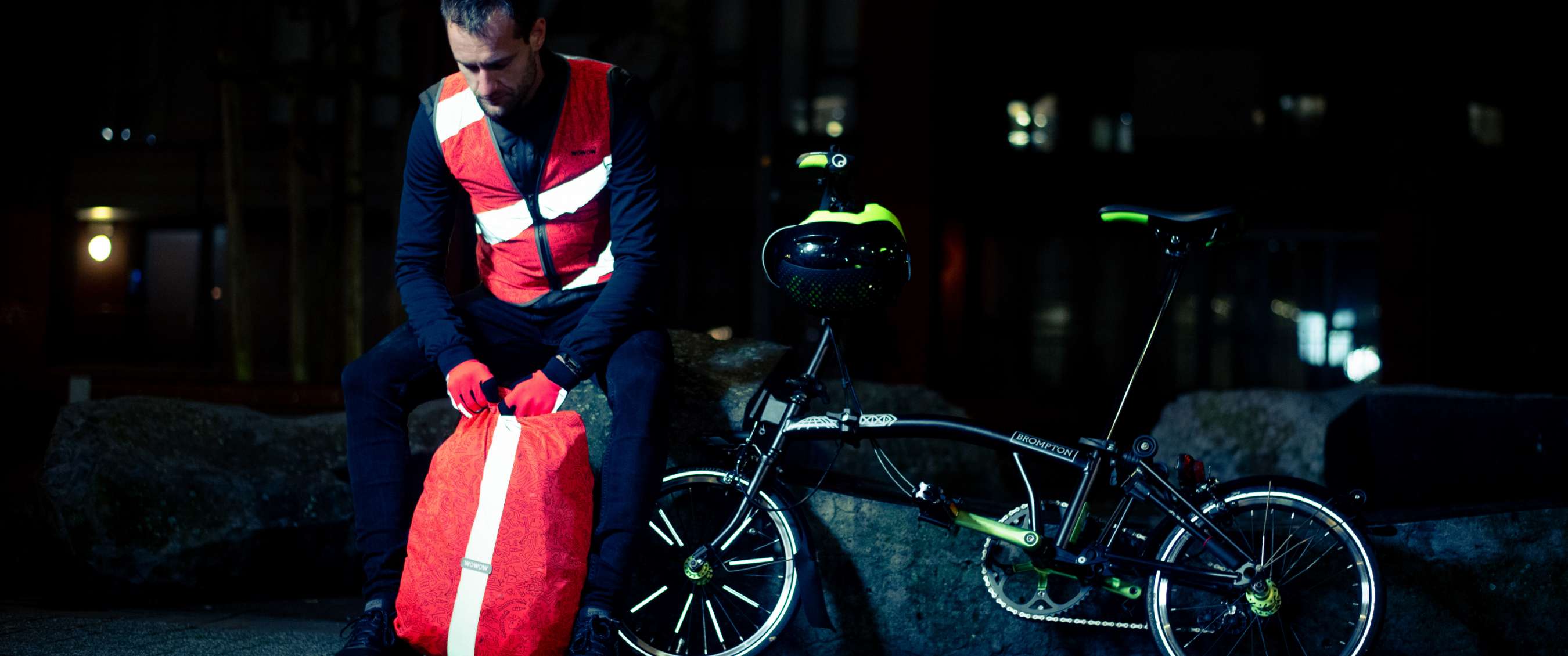 Cycliste avec gilet réfléchissant rouge assis près d’un vélo pliant et tenant un sac réfléchissant.
