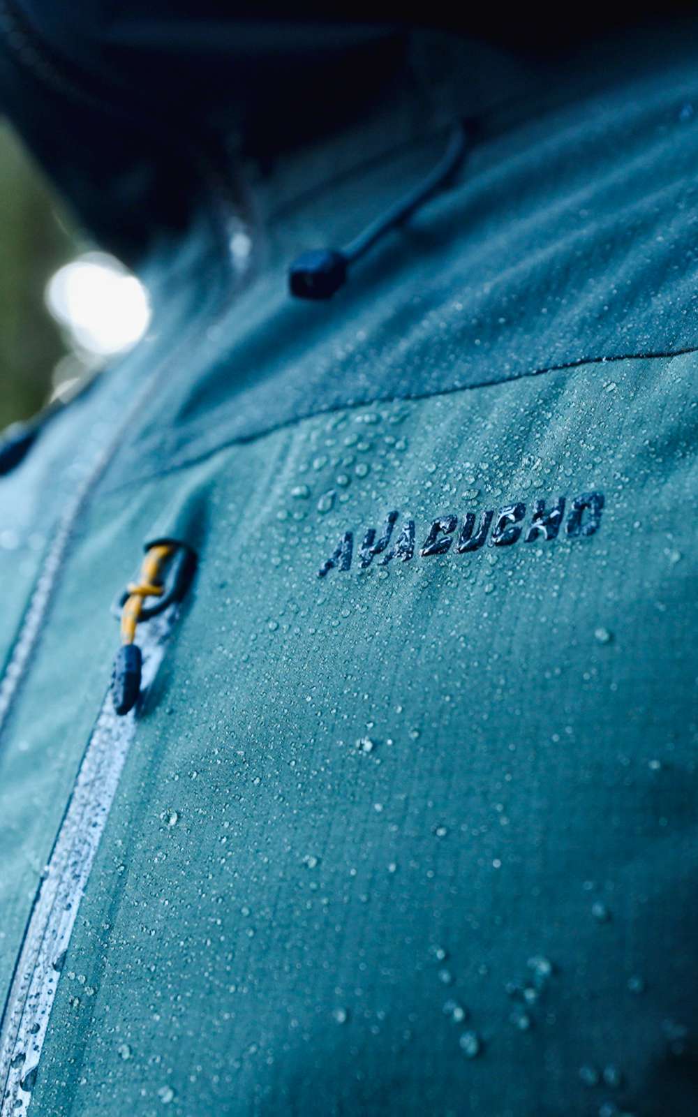 Des gouttes d’eau reposent sur une veste outdoor bleue avec fermeture et logo Ayacucho visible.