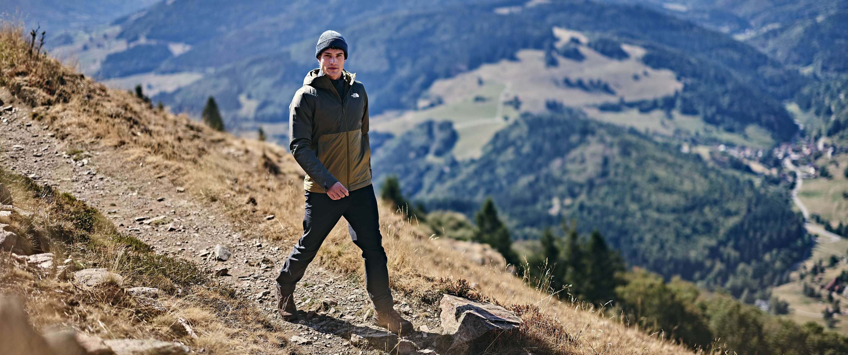 Un randonneur en veste verte et bonnet gris marche sur un sentier de montagne surplombant une vallée.