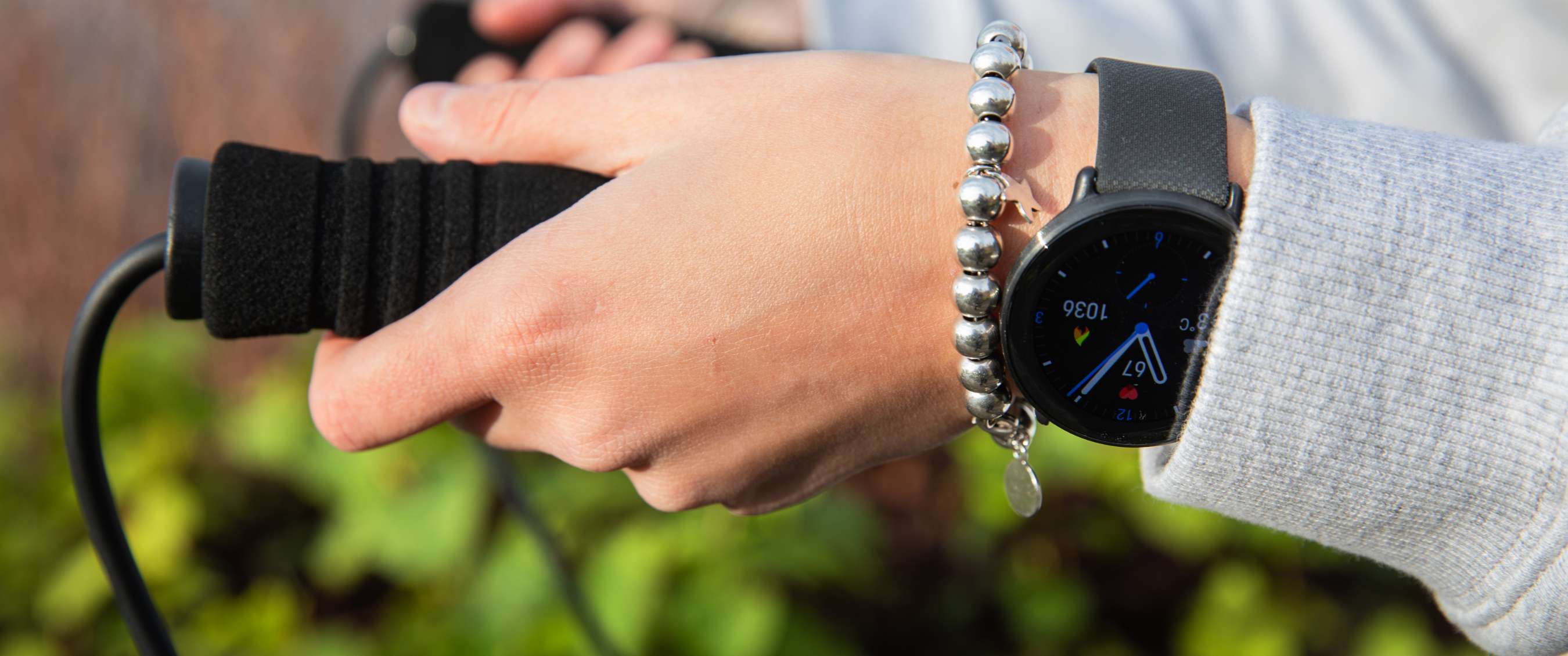 Main portant une montre connectée noire et un bracelet argenté, tenant une poignée noire dehors.