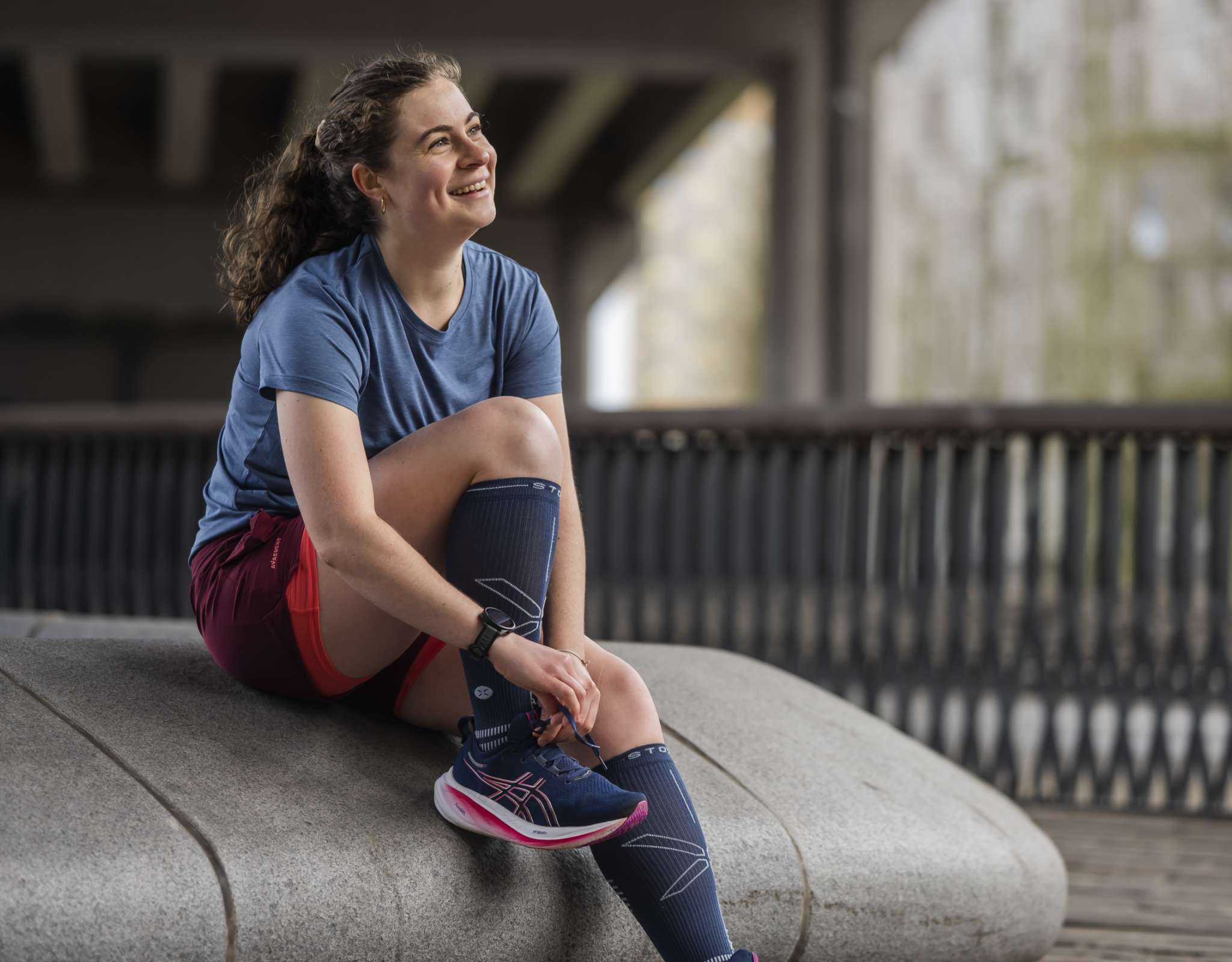 Femme assise sur un banc en pierre, attachant sa chaussure de course, en t-shirt bleu et short bordeaux.