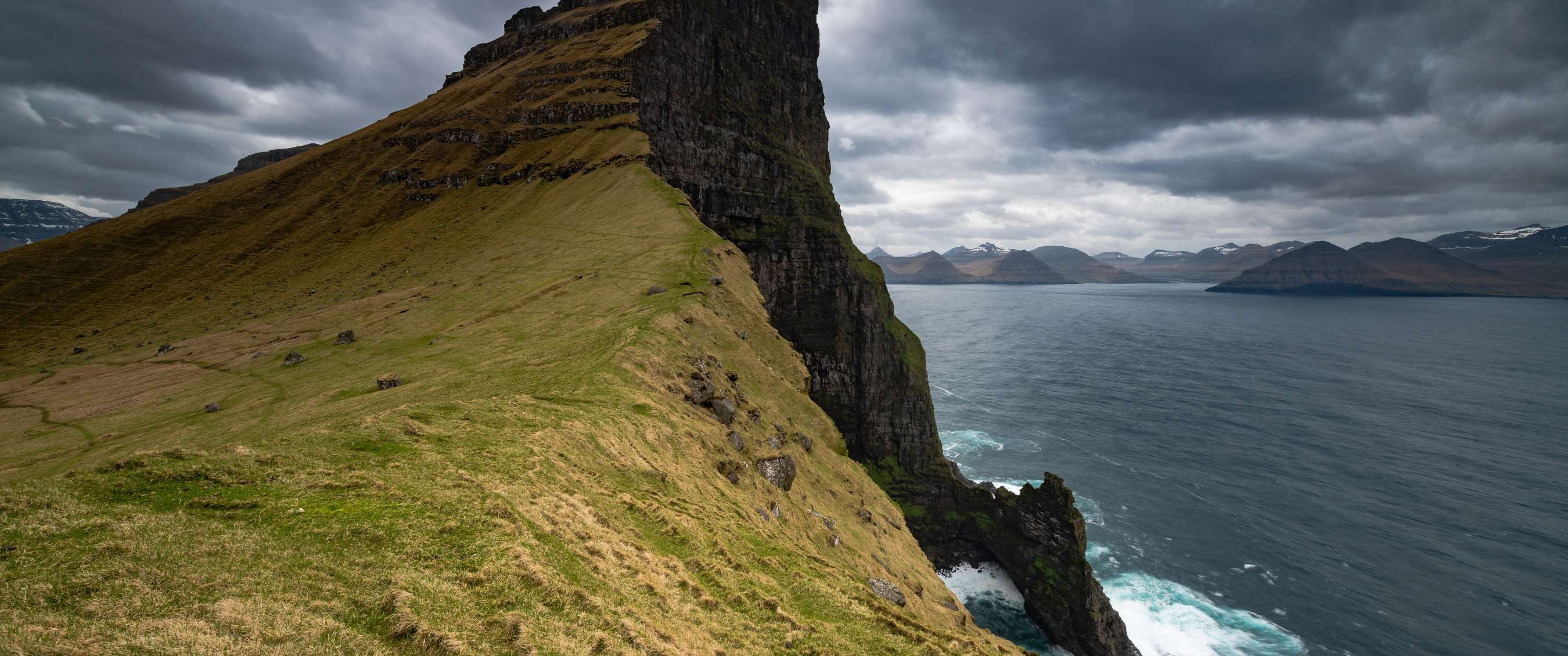 Pente herbeuse raide le long d’une falaise sombre au-dessus de la mer sous un ciel très nuageux.