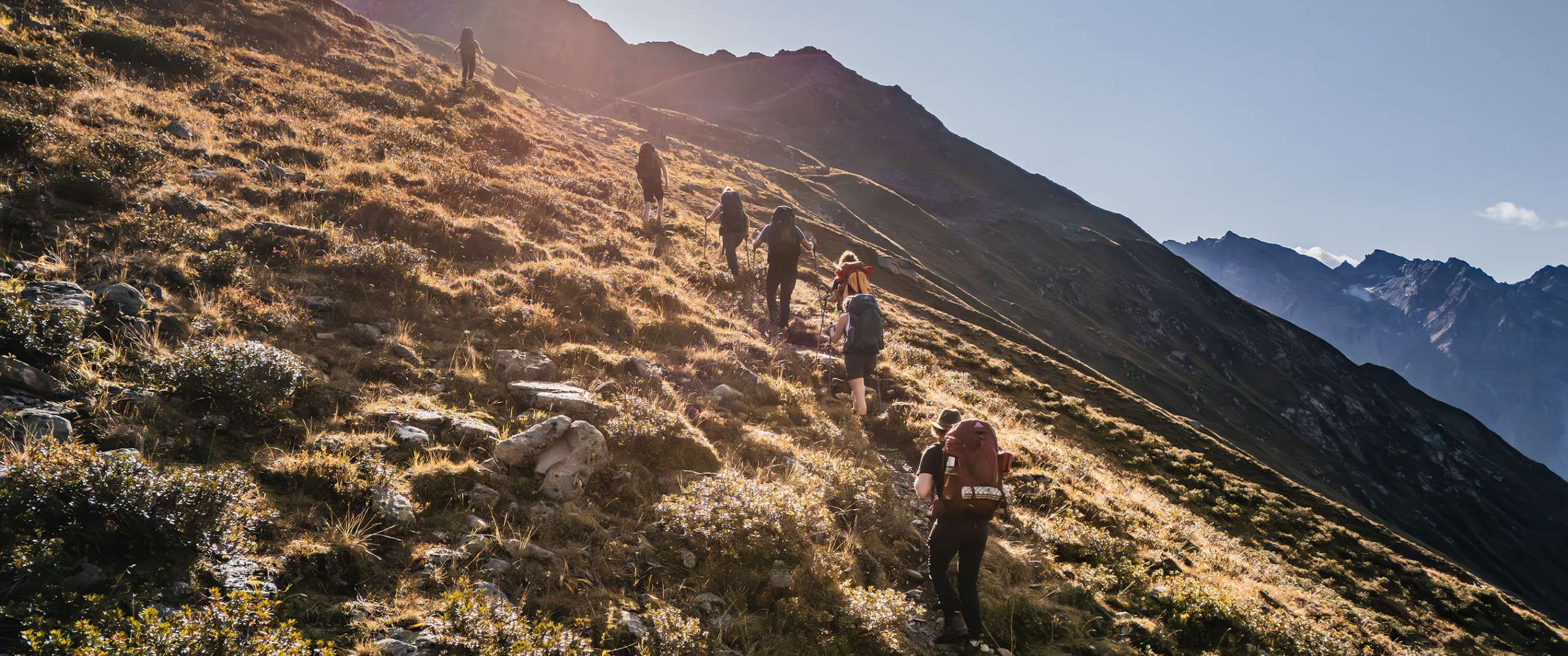 Des randonneurs marchent en file sur une pente de montagne ensoleillée.