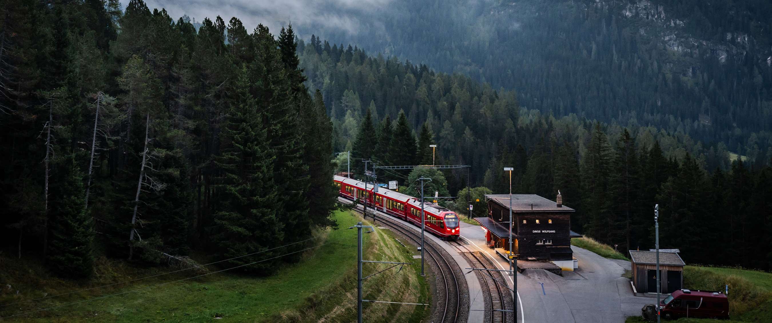 Train à voie étroite passant par une gare de montagne entourée de conifères.