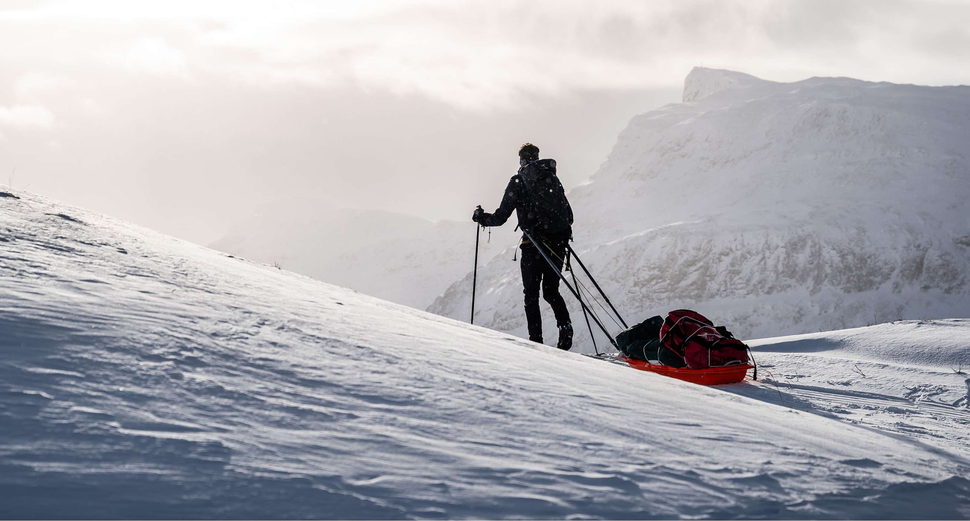 Personne avançant avec des bâtons et un traîneau chargé dans un paysage montagneux blanc.