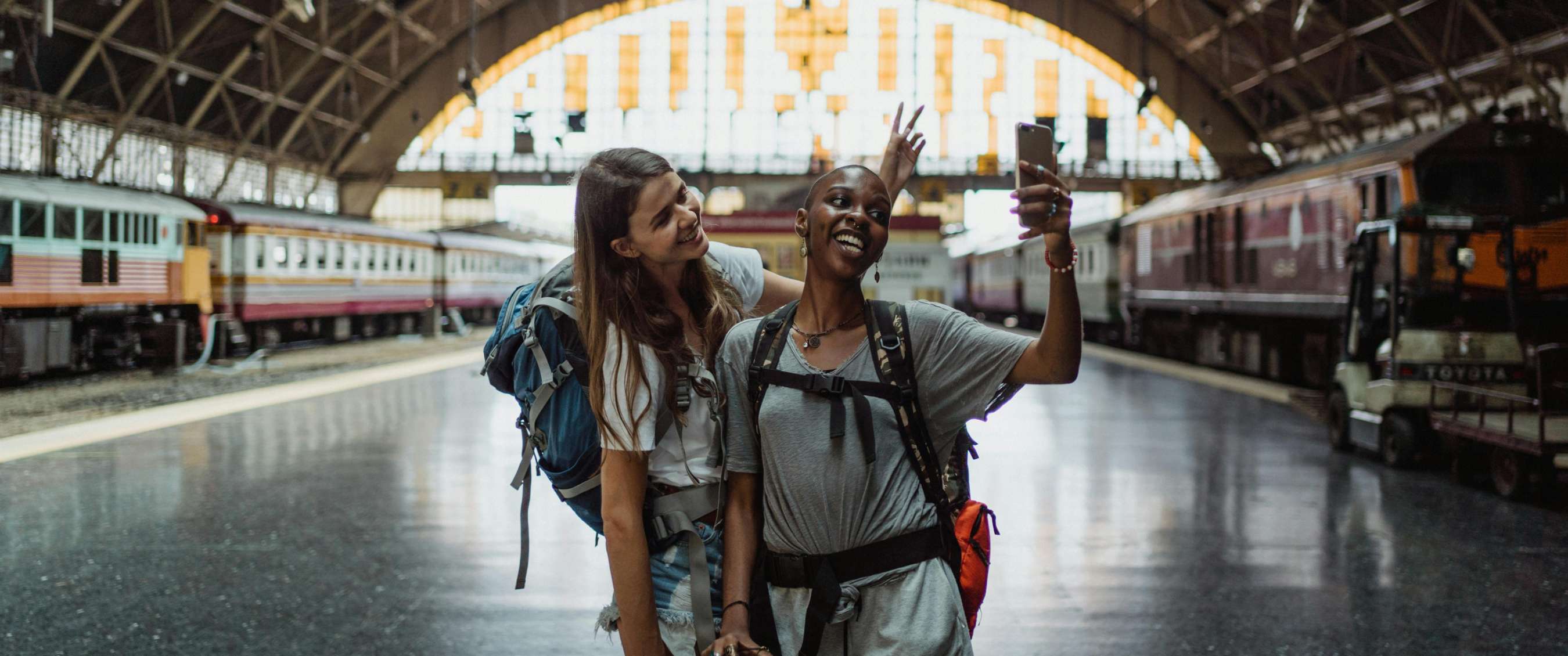 Deux images : deux voyageurs avec sacs à dos prennent un selfie dans une gare entre des trains à l’arrêt ; grande halle de gare avec quais et trains en arrière-plan.