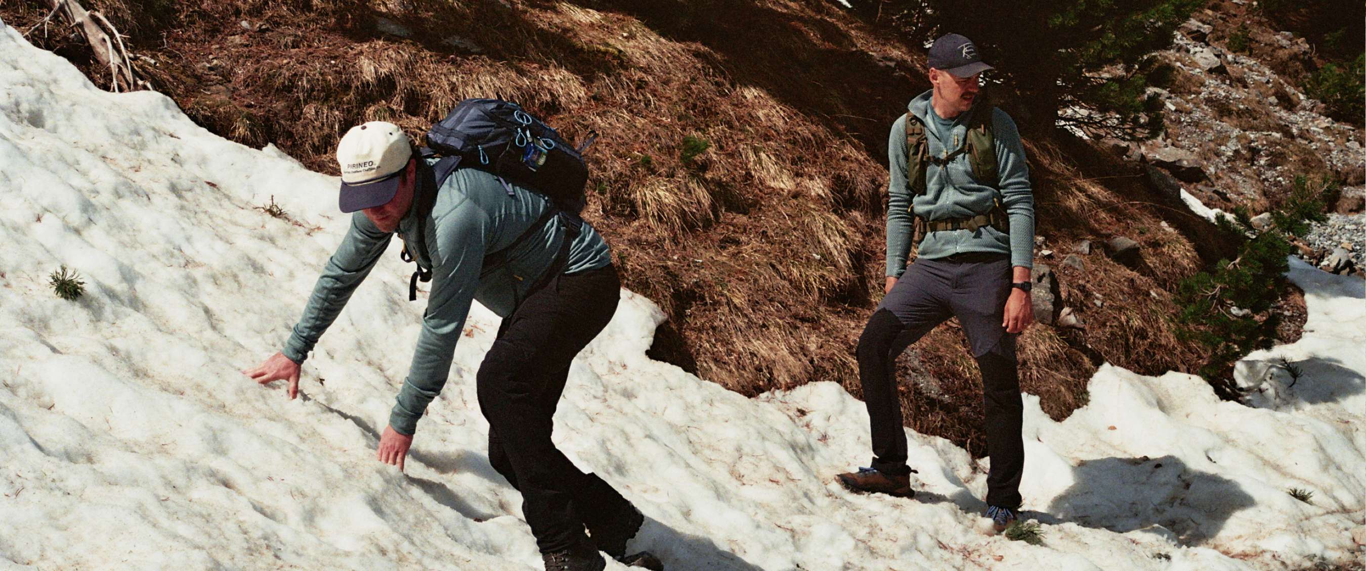Deux randonneurs se tiennent près d’un lac de montagne devant des sommets rocheux enneigés.