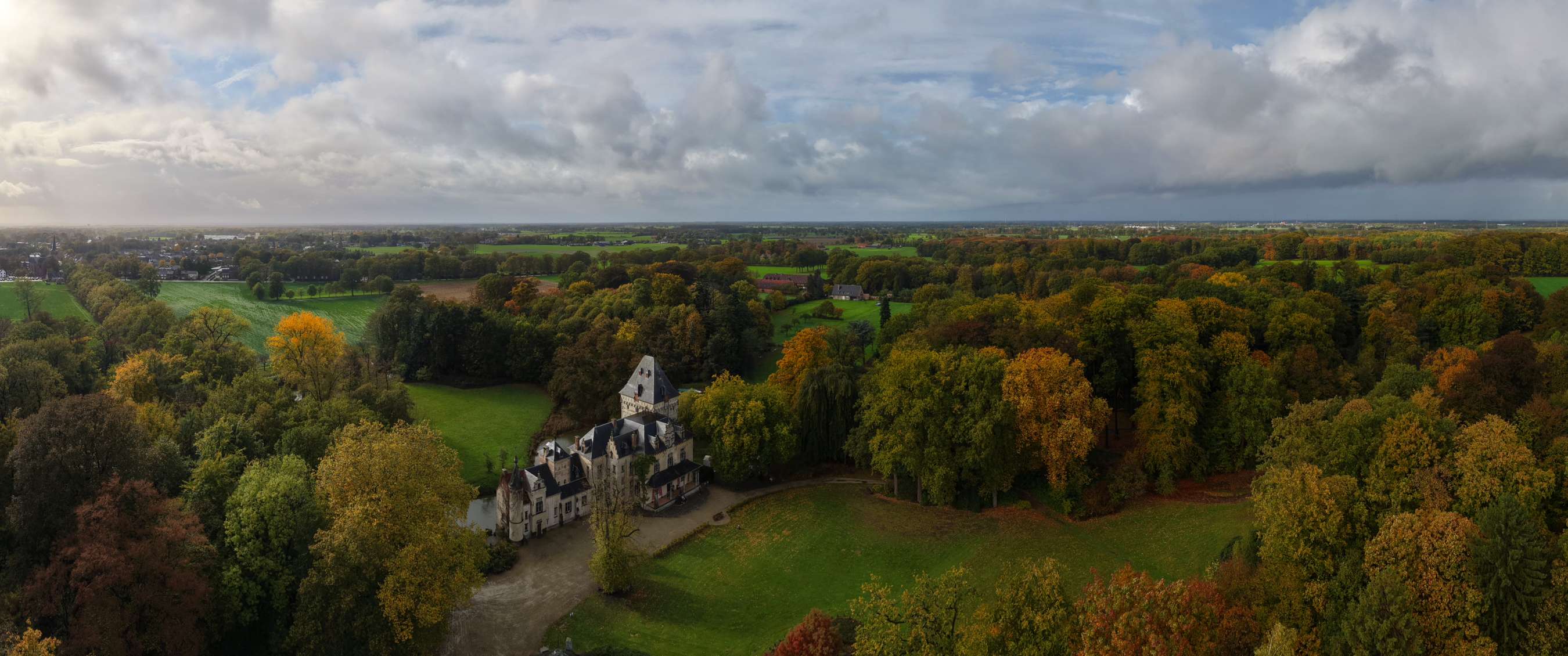 Paysage panoramique avec un château historique bordé d’arbres aux couleurs d’automne et de larges espaces verts.