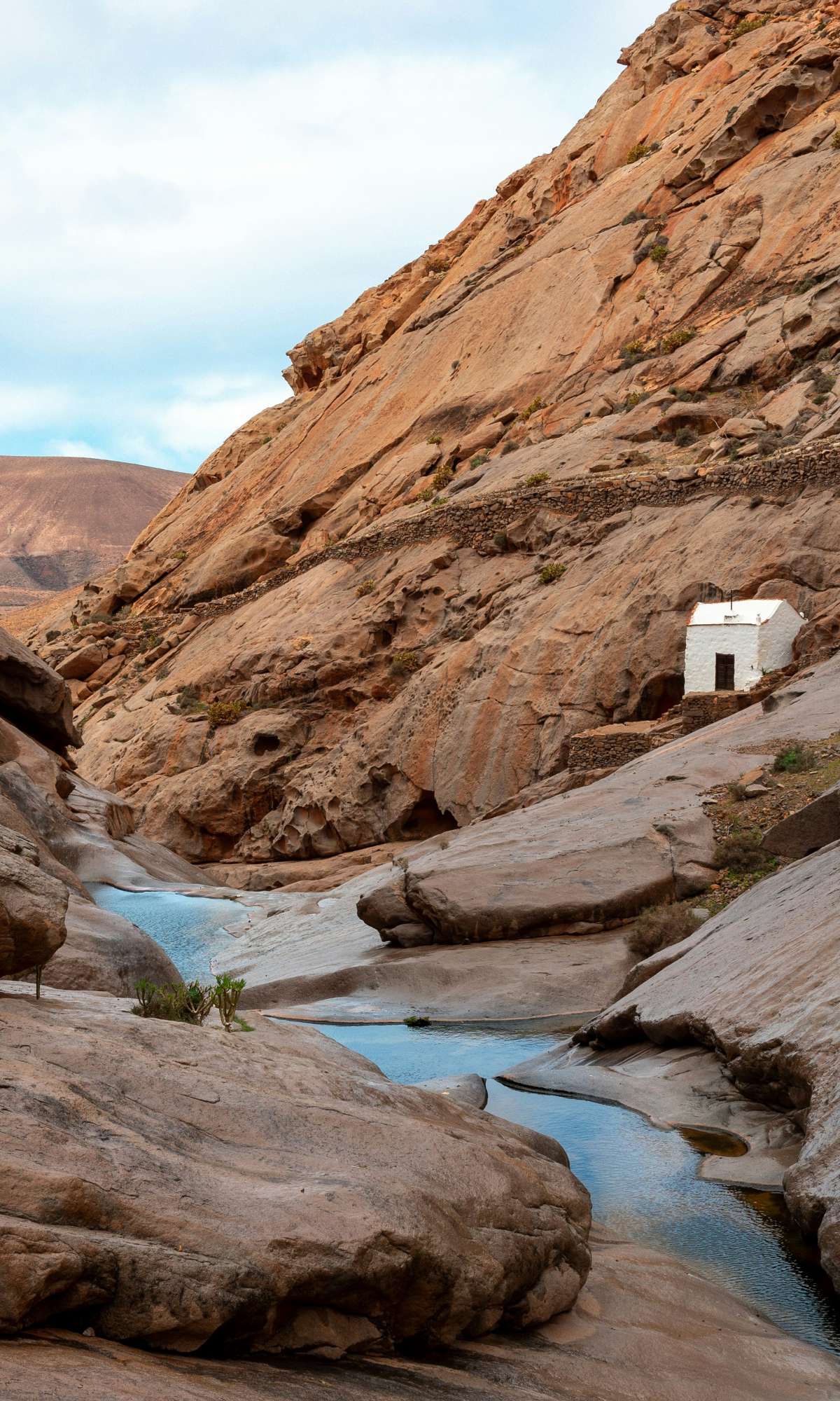 Petit cours d’eau entre des rochers lisses dans un canyon sec avec une maison blanche sur la pente.