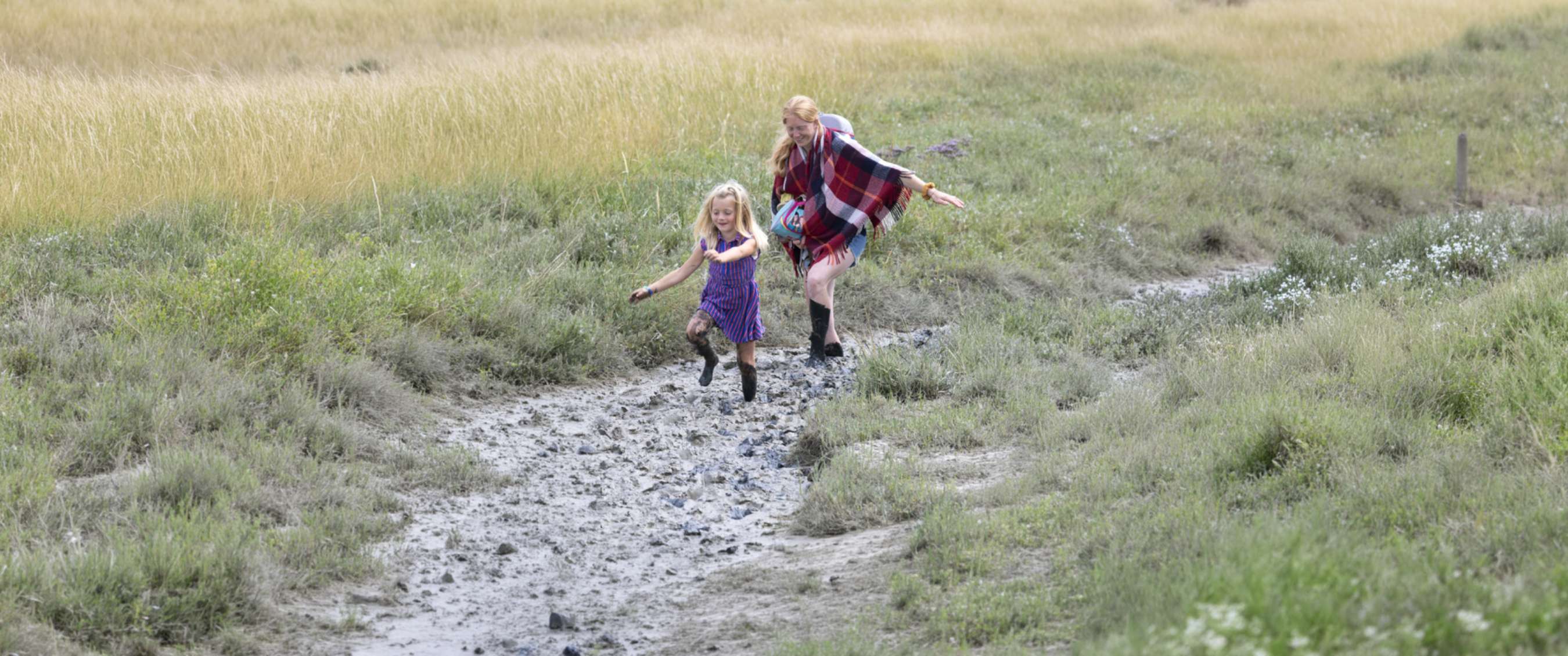 Un enfant et un adulte avancent les jambes boueuses sur un sentier humide dans l’herbe.