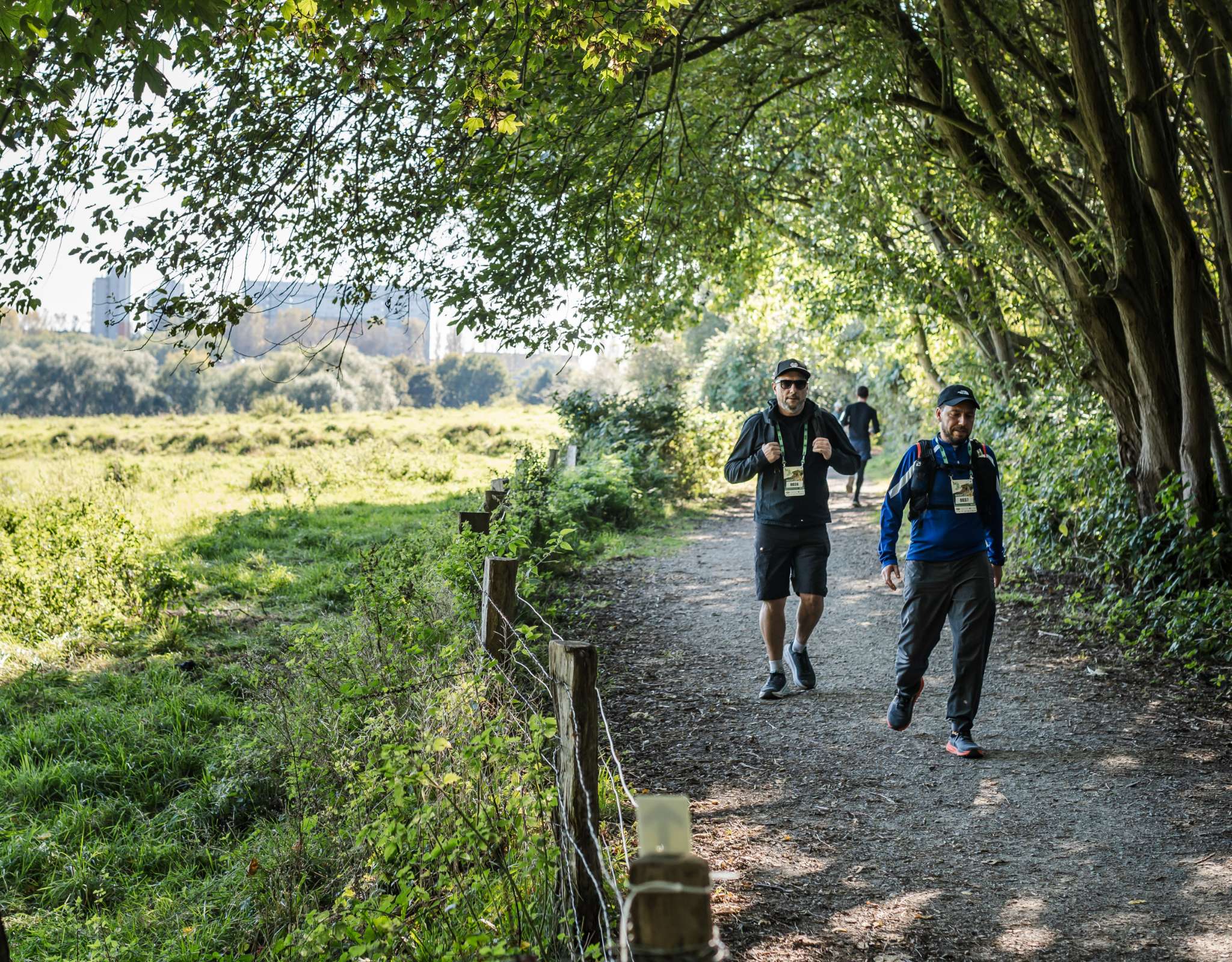Deux randonneurs suivent un sentier ombragé le long de champs verdoyants.