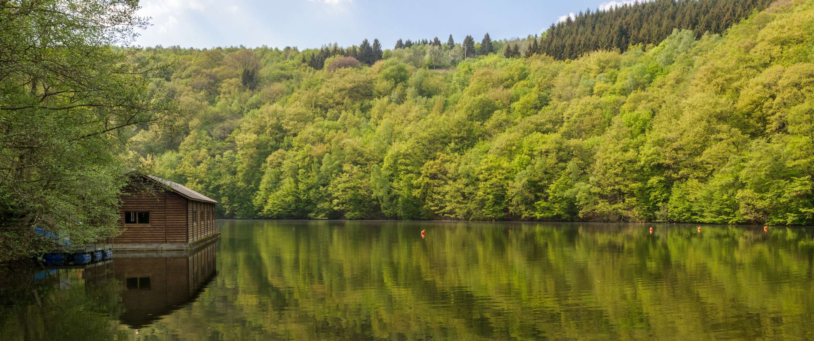 Lac boisé avec bâtiment en bois sur la rive et reflet des arbres dans l’eau.