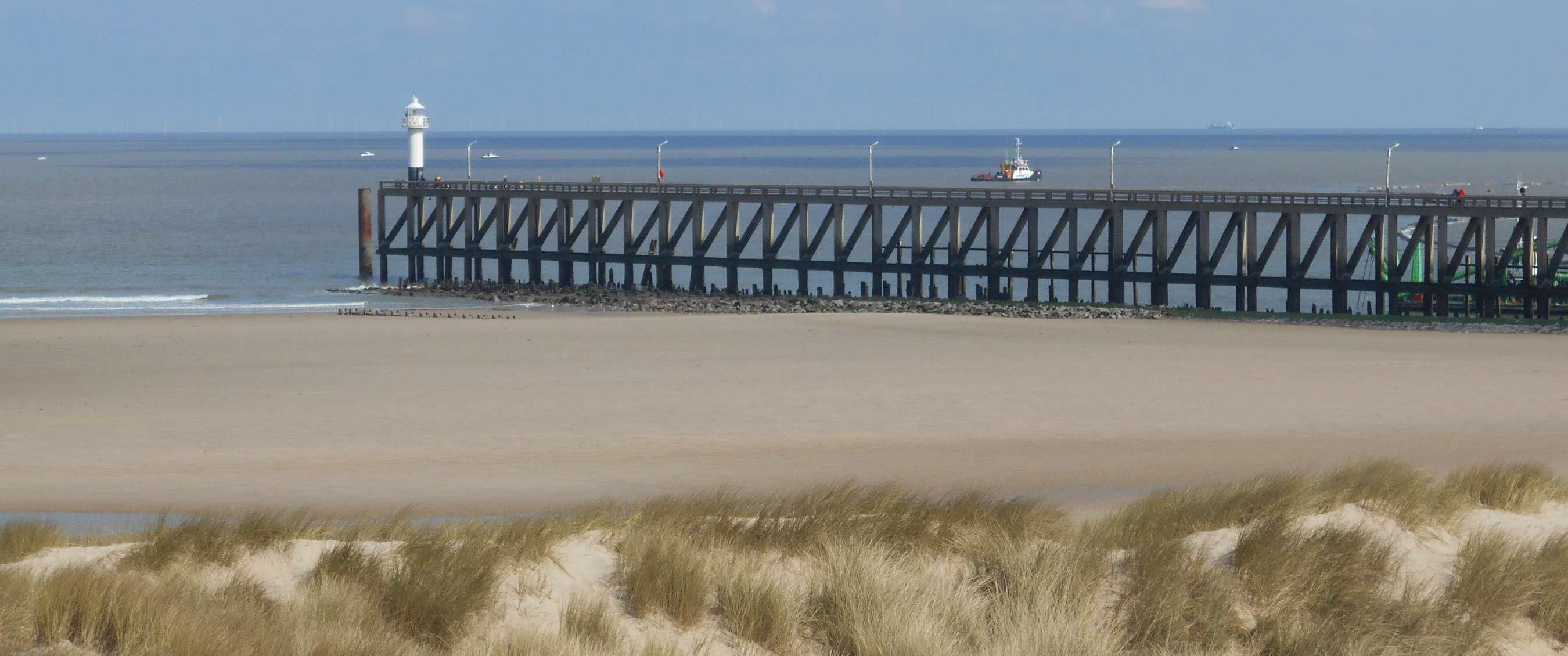 Longue jetée avec un phare s’avançant dans la mer, vue depuis une large plage de sable avec des dunes herbeuses.