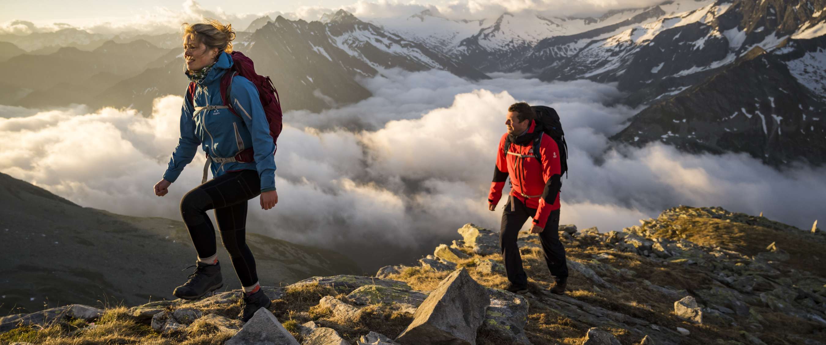 Deux randonneurs montent sur un sentier rocheux au-dessus des nuages et sommets enneigés.