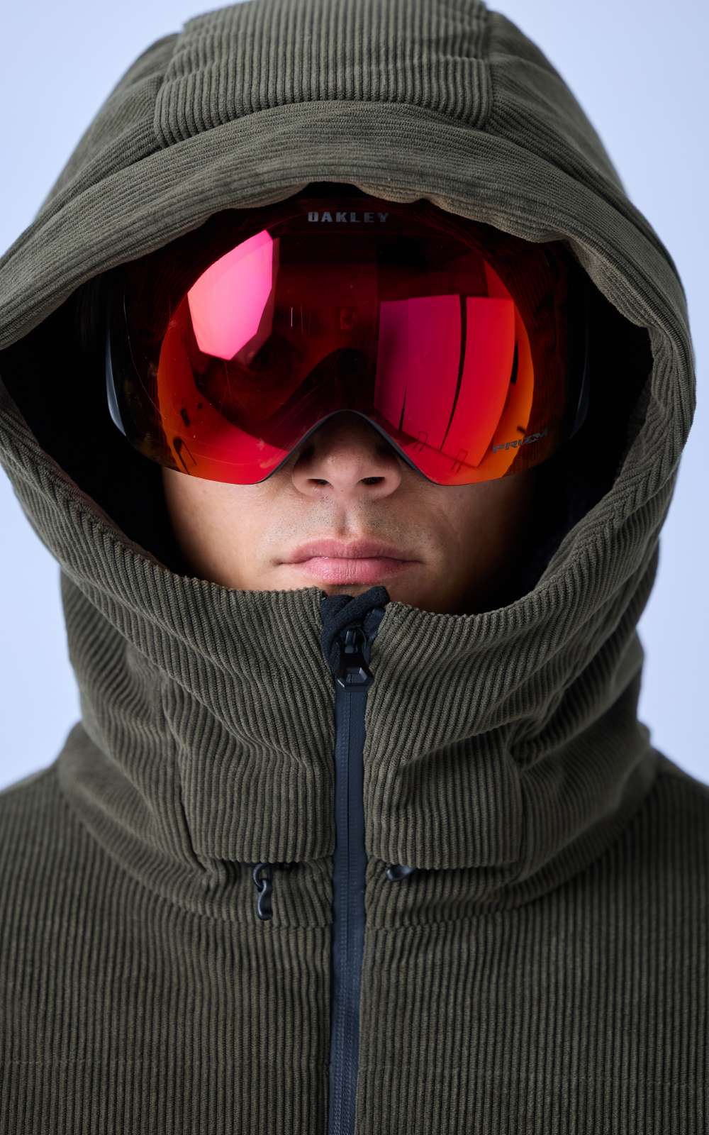 Gros plan d'une personne avec un masque de ski Oakley à écran miroir rouge sous une capuche verte.