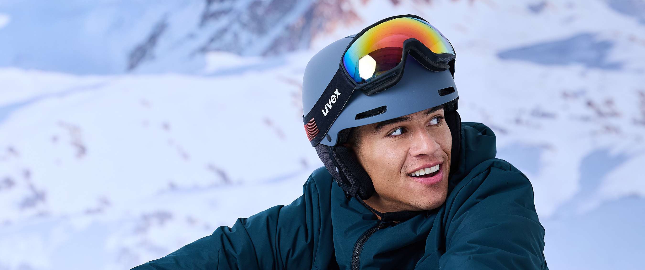 Un homme en veste de ski vert foncé est assis dans la neige, avec un masque Uvex sur son casque.