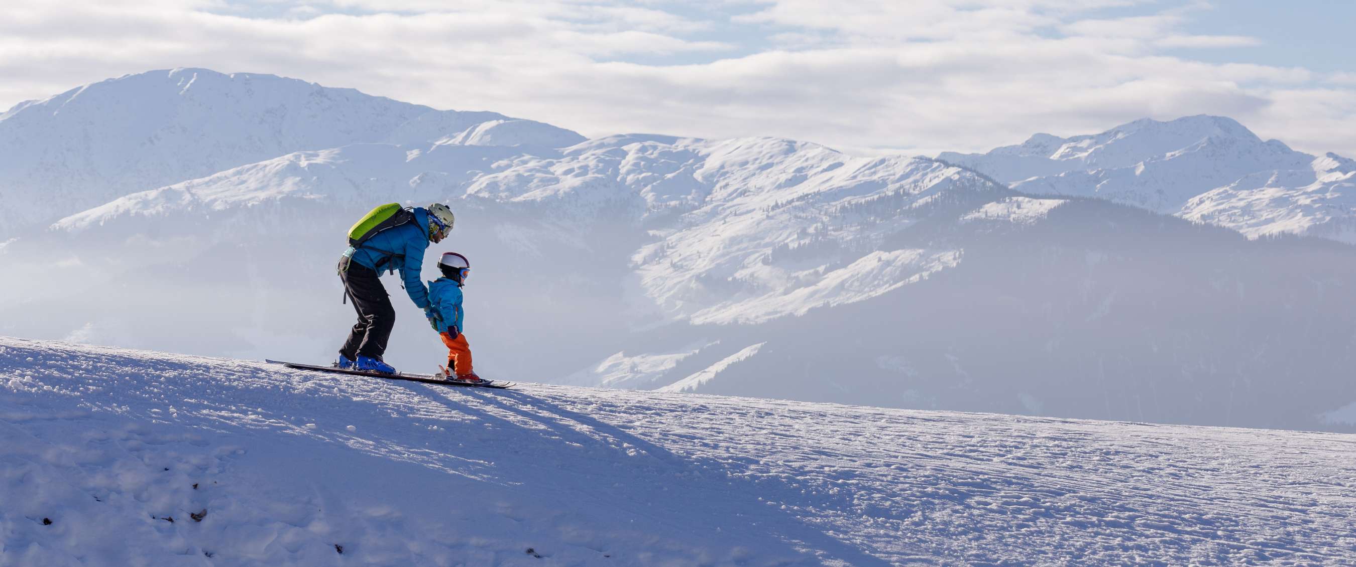 Un adulte aide un enfant à skier sur une pente. Des montagnes enneigées en arrière-plan.