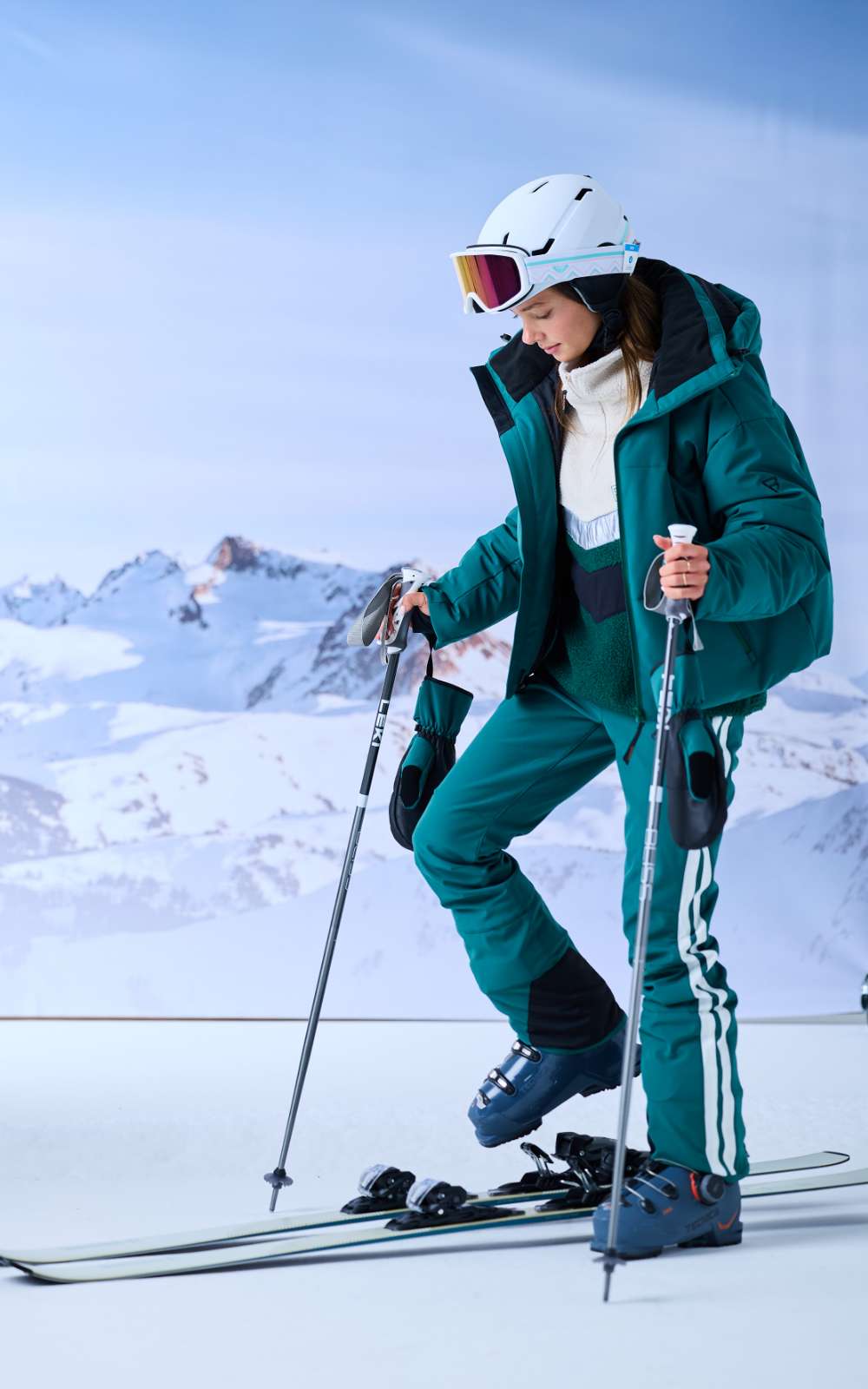 Une skieuse avec casque blanc et masque regarde ses skis devant un décor de montagnes enneigées.