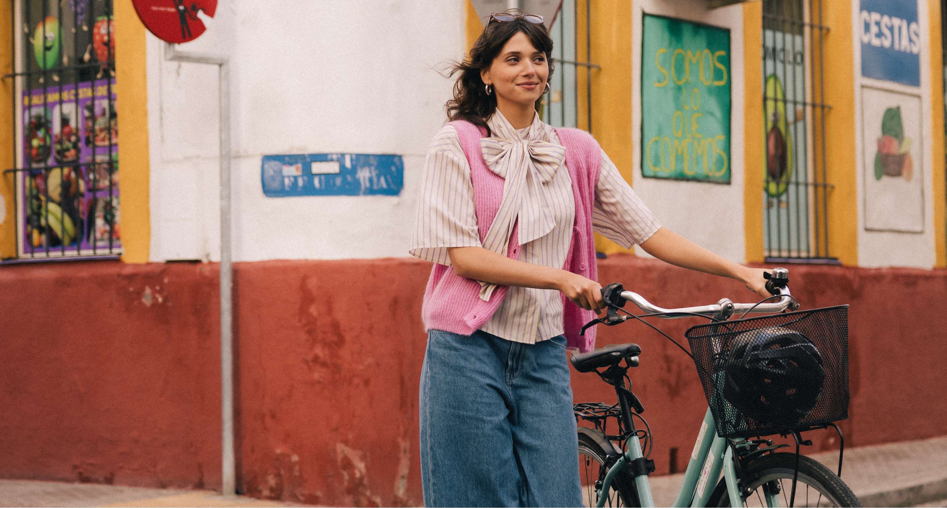 Femme en jean large et gilet rose marchant avec un vélo devant une façade colorée.