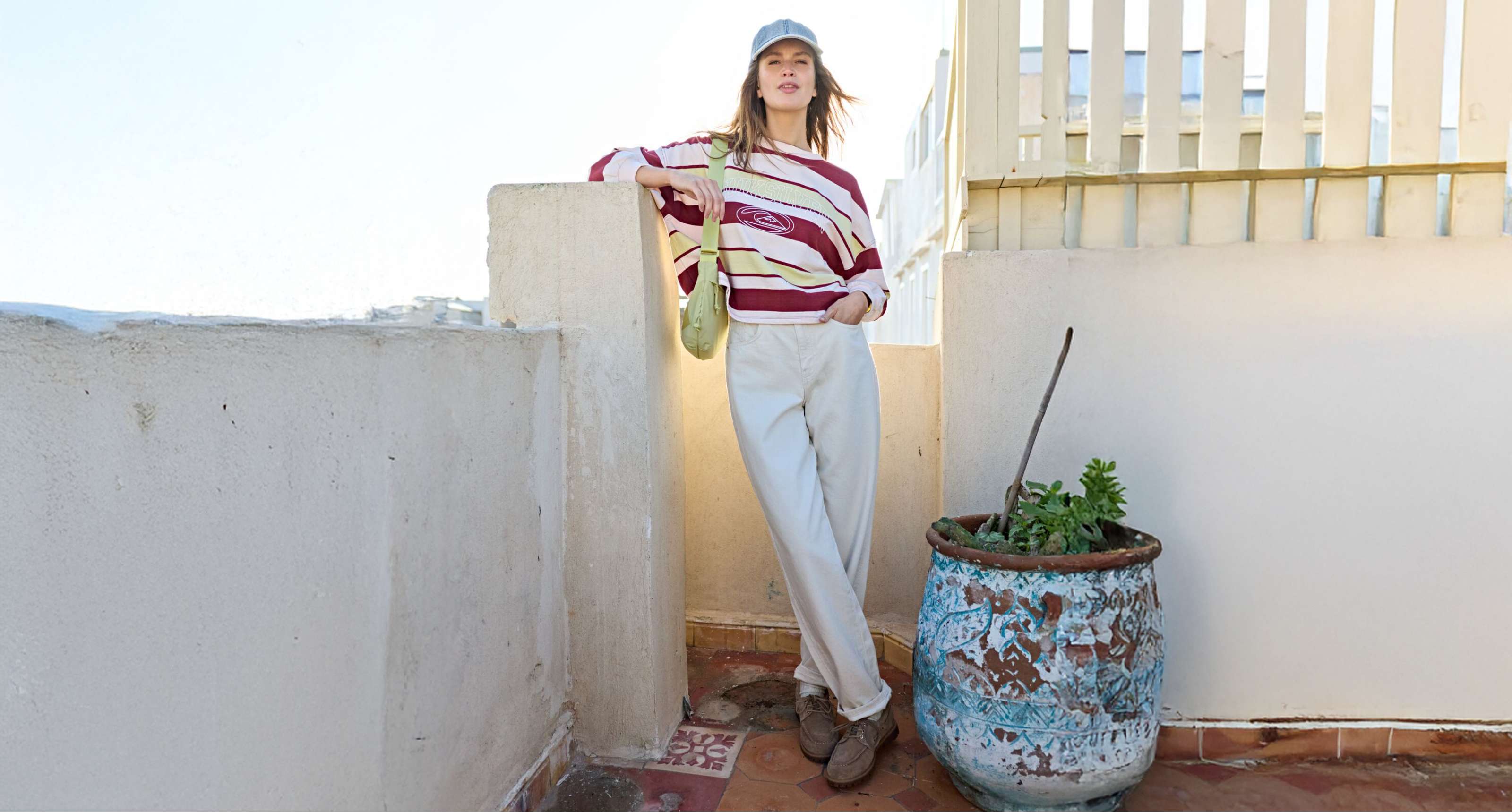 Femme en pantalon large clair et pull rayé appuyée contre un muret sur une terrasse ensoleillée.