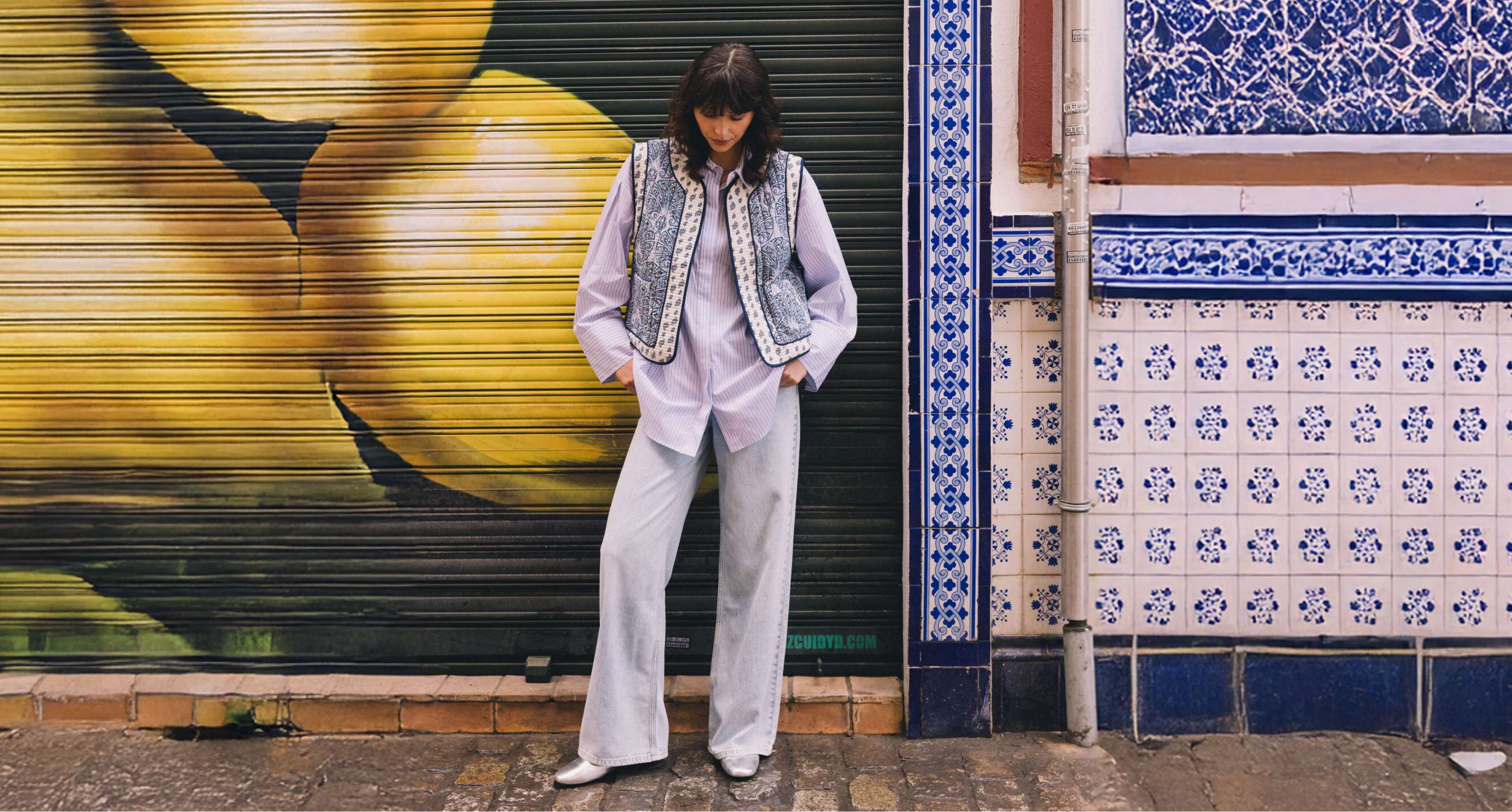 Femme en jean clair, chemise rayée et gilet à motifs devant un volet peint en jaune.